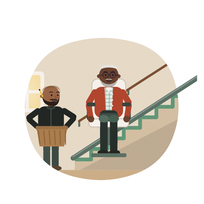 A man tries a stairlift at home, smiling, while his son waits at the foot of the stairs with a laundry basket.